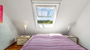 Ein Schlafzimmer mit schrägen Wänden, einem Oberlicht, einem Bett mit gestreifter Bettdecke, einem Nachttisch mit Lampe und einem Beistelltisch ebenfalls mit Lampe.