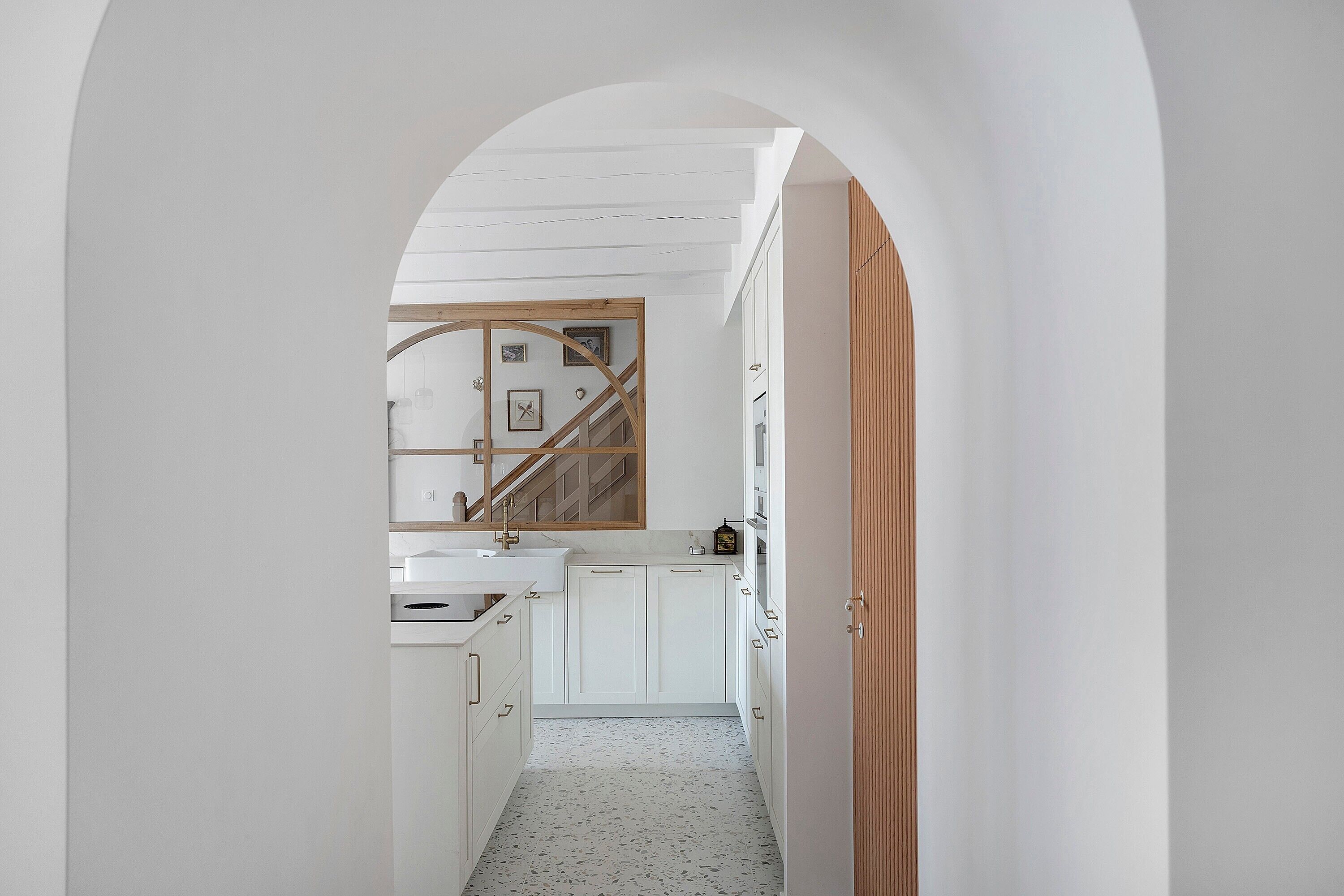 Un couloir étroit mène à une cuisine dotée de meubles blancs, d’un escalier en bois et d’une porte blanche.