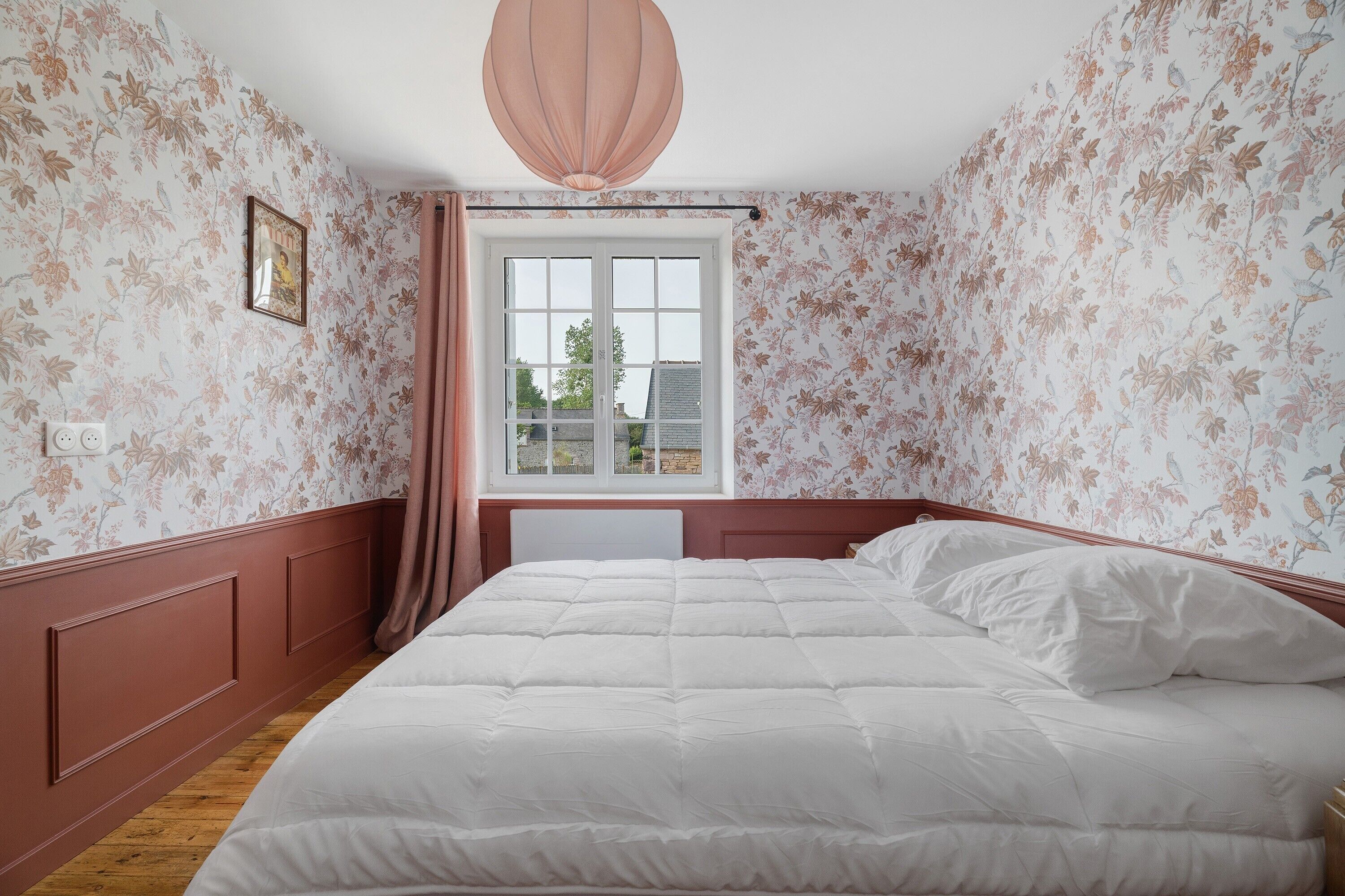 A bedroom with a large bed, a window with curtains, floral wallpaper, and a hanging light fixture.