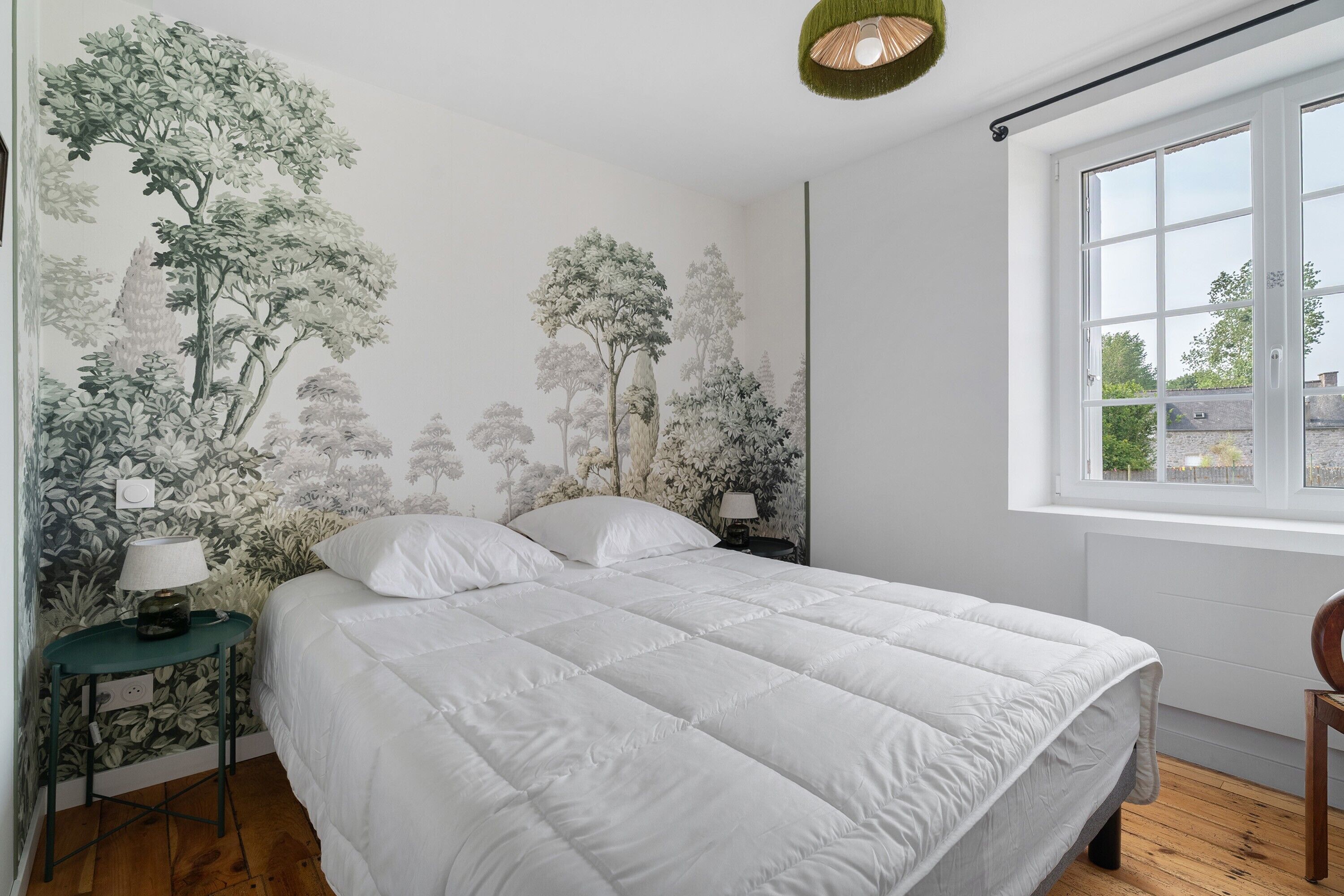 A bedroom with a large bed, a nightstand with a lamp, a window with a view, and a wall with a nature-themed mural.