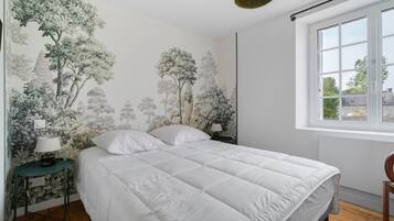 A bedroom with a large bed, a nightstand with a lamp, a window with a view, and a wall with a nature-themed mural.