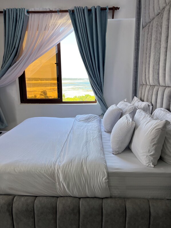 A bed with white bedding, a view of the ocean through a window, and grey curtains. - Luxury Living in Nyali — Wake up to breathtaking ocean view (Mombasa)