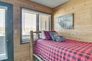 Ein Zimmer in einer Holzhütte mit einem Bett, einem Fenster mit Jalousien und einem Bild an der Wand.