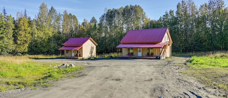 Deux cabanes aux toits rouges, un chemin de terre et un paysage boisé en arrière-plan.