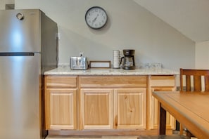 A kitchen with wooden cabinets, a stainless steel refrigerator, a clock, a toaster, a coffee maker, and a wooden dining table. - Kenai River Base! Spacious 2-unit Cabin Compound (Soldotna)