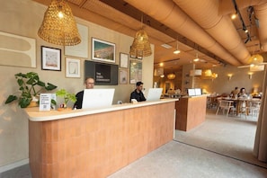 A reception area with two receptionists, a wooden reception desk, framed artwork on the wall, and a dining area in the background. - THE BABEL COMMUNITY Hôtel SQY Gare (Montigny-le-Bretonneux)