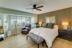 Ein Schlafzimmer mit Bett, Deckenventilator, Sitzbank, Stuhl, Nachttisch mit Lampen und Wandbild.