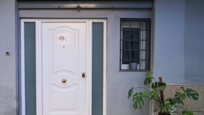Una porta bianca con maniglia in ottone, una piccola finestra con sbarre metalliche e una pianta in vaso sulla destra.
