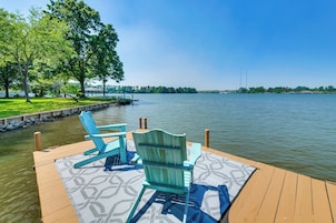Un muelle de madera con dos sillas Adirondack y un tapete estampado, con vista a un lago tranquilo y una orilla lejana.