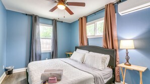 Ein Schlafzimmer mit einem Bett, einem Deckenventilator, einem Nachttisch mit Lampe und einem Fenster mit Vorhängen.