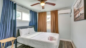 Un dormitorio con cama, ventilador de techo, ventana, cortinas, mesita de noche y un cuadro en la pared.