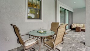 Un patio con una mesa redonda de vidrio, sillas de mimbre, un sofá acolchado y una ventana con cortinas.