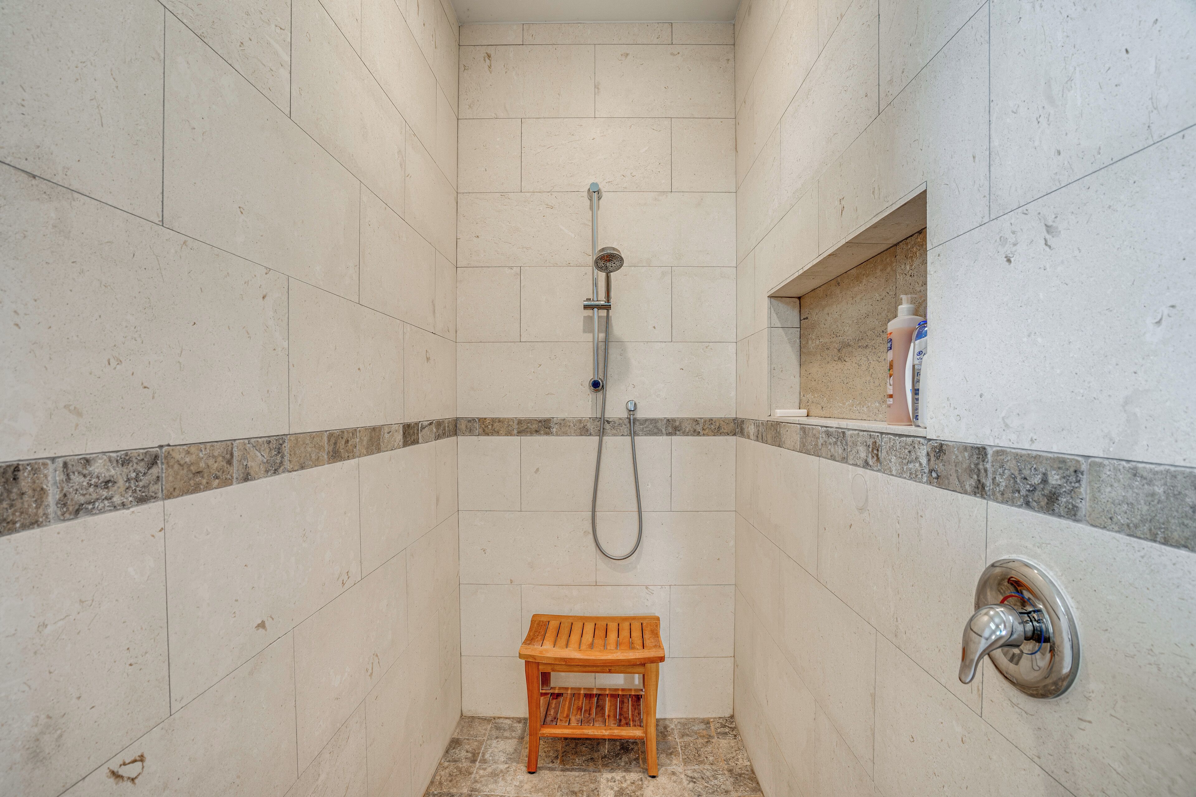 Un espacio de ducha pequeño con un taburete de madera, un cabezal de ducha manual y un grifo empotrado en la pared.