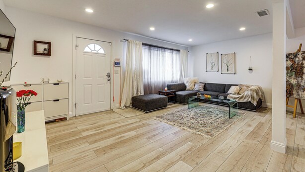 Una sala de estar moderna con un sofá, un reposapiés, una mesa de centro y una alfombra. Hay una puerta, un ventanal con cortinas y una obra de arte colgada en la pared.