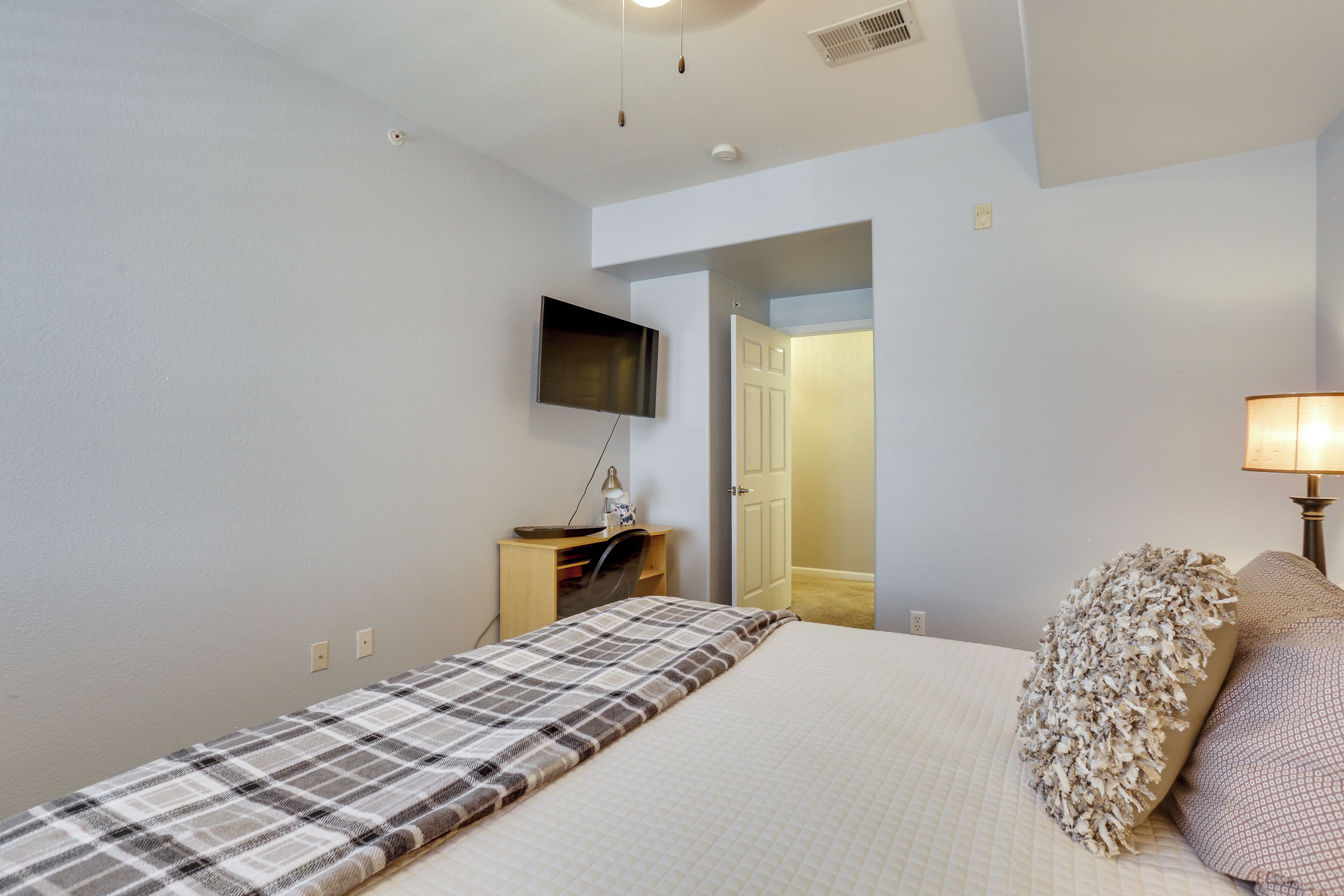 A bedroom with a bed, a television mounted on the wall, a desk with a chair, and a door leading to another room.