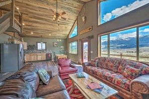 Ein geräumiger Wohnbereich mit großem Fenster und Blick auf die Berge, eine gemütliche Sitzecke mit gemustertem Sofa und einer Holzzahlende mit Deckenventilator.