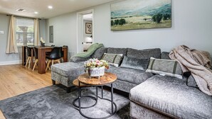 A living room with a grey sofa, a round coffee table with a flower vase, a dining area with a wooden table and chairs, and a painting on the wall.