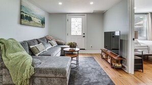 A living room with a grey sofa, a green throw blanket, a small round table with a vase, and a television on a stand.