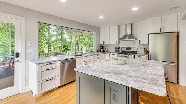 A modern kitchen with white cabinetry, a large island with a marble countertop, and a view of greenery outside.