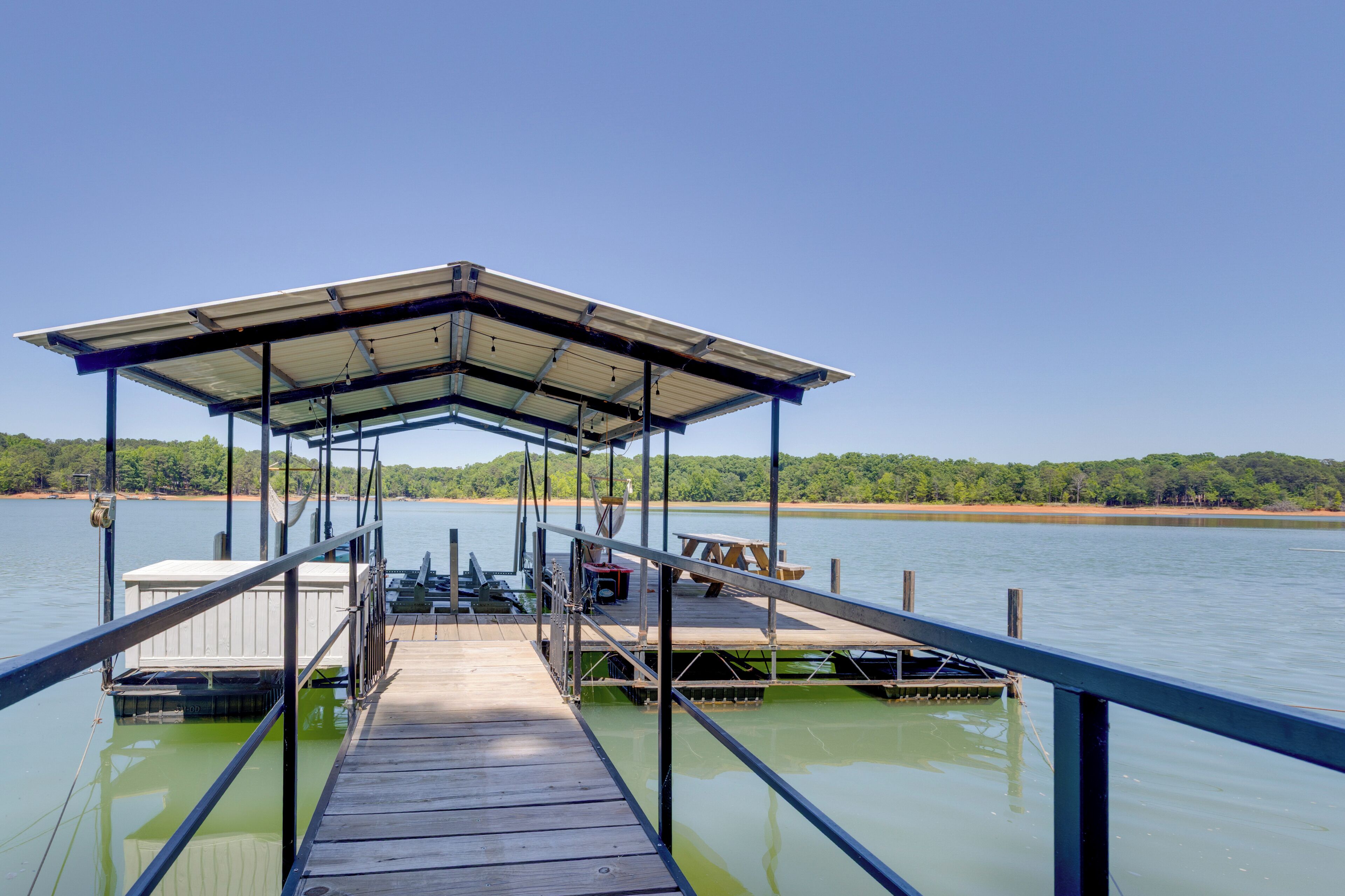 A wooden dock with a covered seating area, a boat, and a view of a calm lake with a forested shoreline.