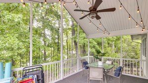 A covered outdoor area with a ceiling fan, string lights, a table with chairs, and a view of a forest.