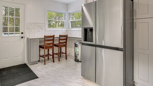 A kitchen with a stainless steel refrigerator, two wooden chairs, a trash can, and a window with a view of trees. - Deck & Patio! Wurtsboro Haven Near Lakes & Trails (Wurtsboro)