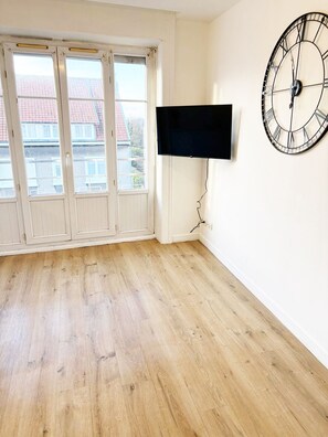 Une pièce dotée d’un plancher en bois, d’une grande fenêtre avec des volets blancs et d’un téléviseur fixé au mur.