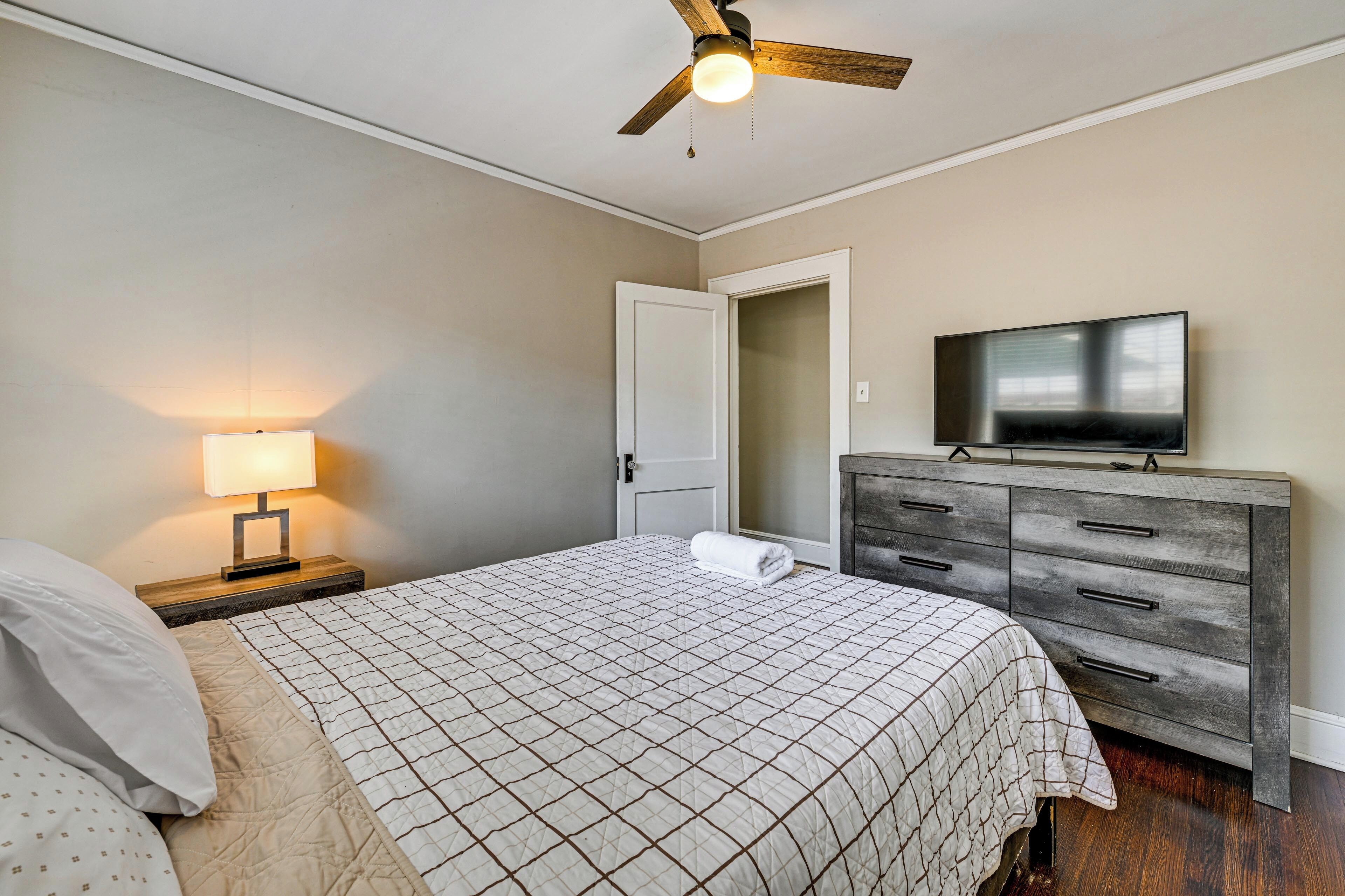 A bedroom with a bed, a nightstand with a lamp, a dresser, a television, and a ceiling fan.