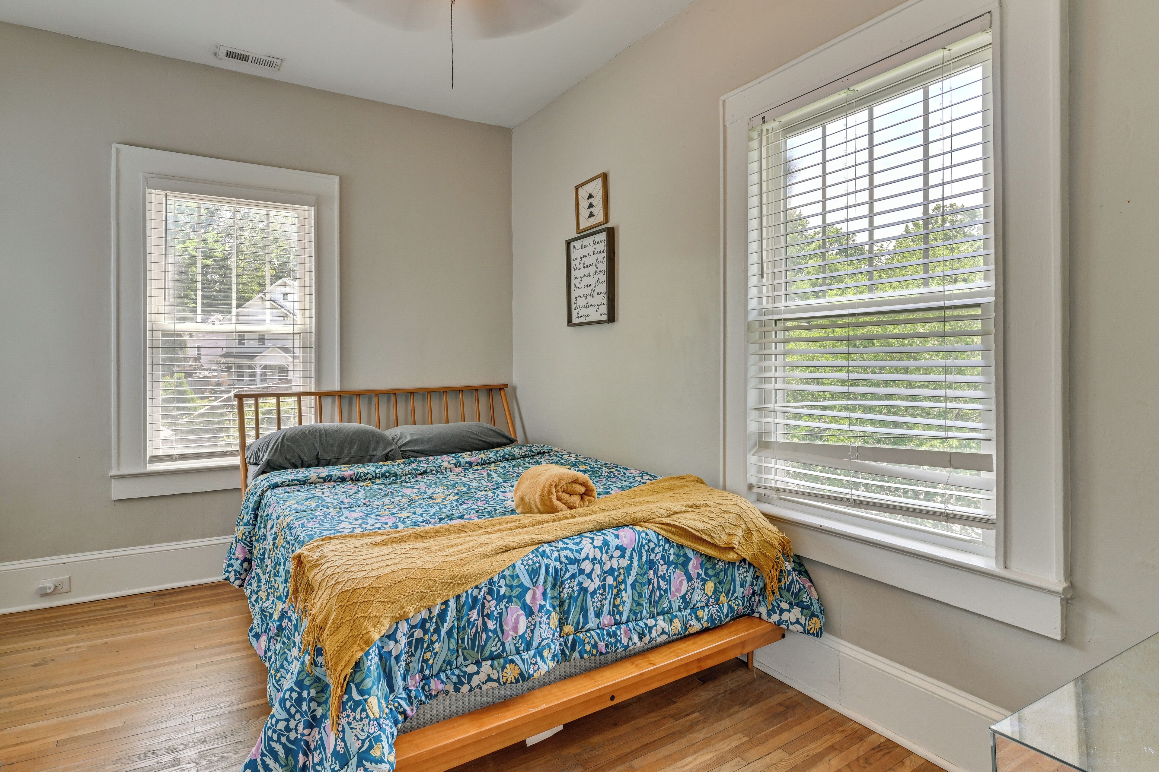 A bedroom with a wooden bed, a window with blinds, a wall-mounted picture, and a view of houses outside.
