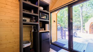 A compact kitchen with a built-in refrigerator, shelves with various items, and a sliding glass door leading to an outdoor deck. - Glamping at Four Fillies Lodge-The Maid Marian (Peterstown)