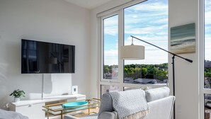 Ein modernes Wohnzimmer mit Flachbildfernseher, einer Couch, einem Couchtisch und einem großen Fenster mit Blick auf die Stadt.
