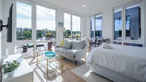 Ein modernes Hotelzimmer mit großen Fenstern, einem Bett, einer Couch, einem kleinen Tisch und einem Fernseher.