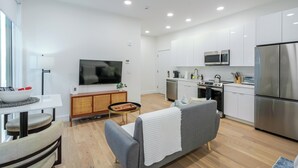 Eine moderne Küche mit weißen Schränken, einem großen Flachbildfernseher, einer Essecke mit Tisch und Stühlen sowie einem grauen Sofa.