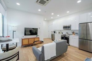 Eine moderne Küche mit weißen Schränken, einem großen Flachbildfernseher, einer Essecke mit Tisch und Stühlen sowie einem grauen Sofa.