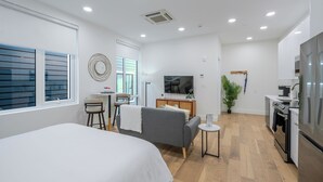 Ein modernes, helles Zimmer mit einem Bett, einem grauen Sofa, einem kleinen Esstisch, einem Fernseher und einer Küchenzeile.