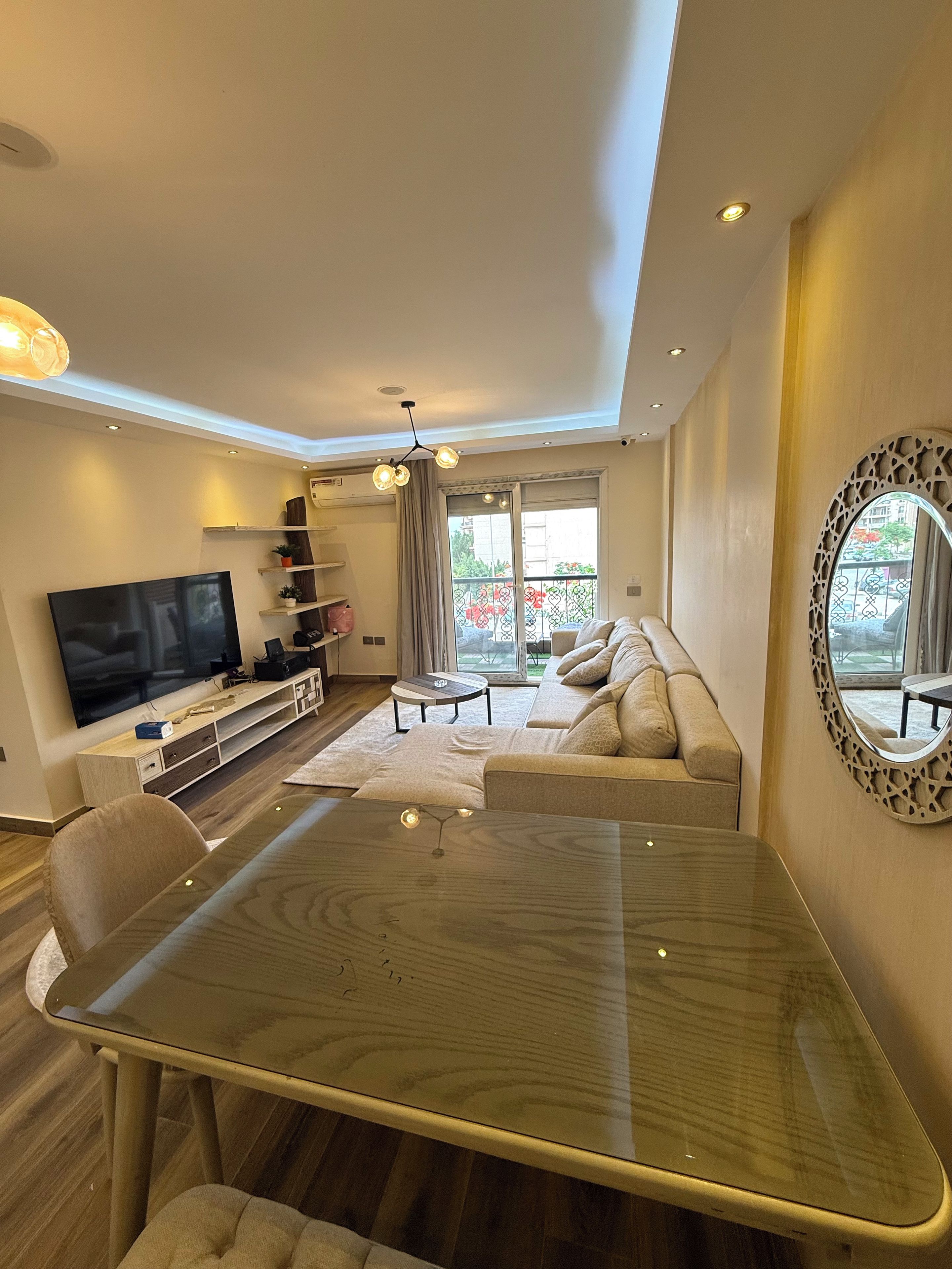 A modern living room with a flat-screen TV, a glass-top dining table, a sofa, and a balcony with a view.