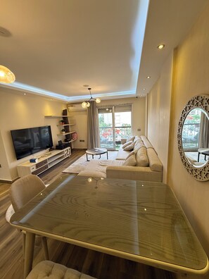 A modern living room with a flat-screen TV, a glass-top dining table, a sofa, and a balcony with a view. - Jacuzzi & Balcony — Rehab Apt Near Top Malls&Dining (Cairo Governorate)