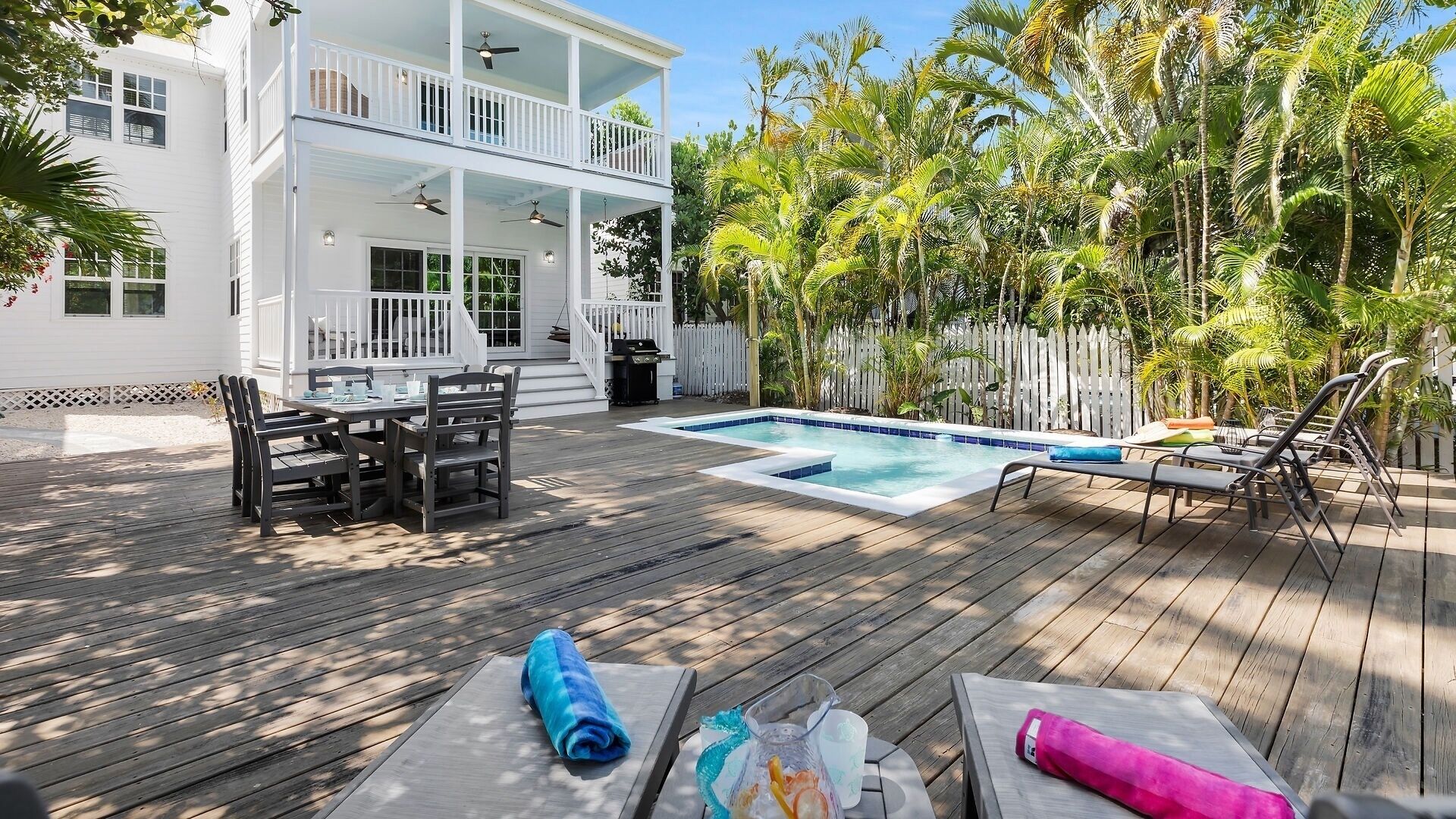 Une terrasse en bois avec une piscine, du mobilier d’extérieur et un bâtiment blanc doté de balcons.