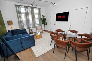 A modern living room with a blue sofa, brown leather chairs, a glass coffee table, and a TV displaying 'NETFLIX'.