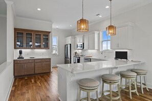 A modern kitchen with white cabinetry, a large island with stools, and wooden flooring. - Oceanside Home with Elevator in Atlantic Beach, NC! (Once Upon a Tide) (Atlantic Beach)