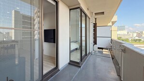 A balcony with a city view, sliding glass doors, a flat-screen TV, and a metal railing. - Lafort Honkawa (Hiroshima City)