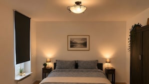 Una camera da letto con un letto, due lampade sui comodini, una finestra con una pianta, un termosifone e un quadro appeso al muro.