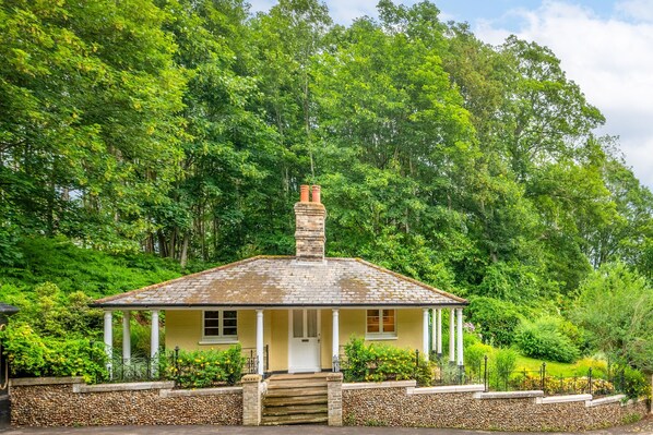 A single-story yellow house with a white porch, a chimney, and a stone wall surrounding the property.