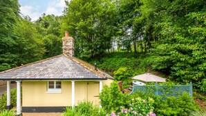 Una casa amarilla con chimenea, rodeada de abundante vegetación y un jardín con flores rosadas.
