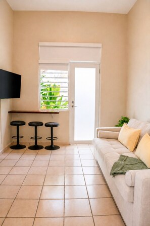 Ein Wohnzimmer mit beiger Couch, drei schwarzen Hocker, einem flachbildfernseher an der Wand und einer Glastür zum Balkon mit Grünpflanzen.