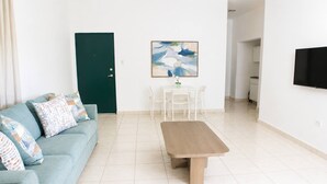Ein Wohnzimmer mit einer Couch, einem Couchtisch, einem Esstisch mit Stühlen, einer grünen Tür, einem Bild an der Wand und einem an der Wand montierten Fernseher.