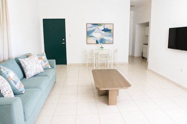 A living room with a sofa, a coffee table, a dining table with chairs, a green door, a painting on the wall, and a TV mounted on the wall. - Spacious 1BR Apt Steps from Beach and Old San Juan (San Juan)