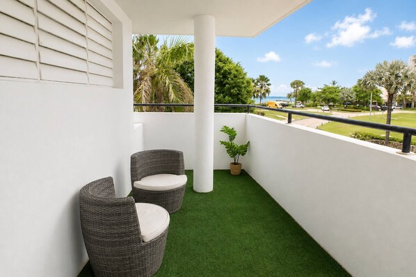 Un balcone con erba sintetica, due sedie in vimini, una pianta in vaso e vista sulla spiaggia e sulle palme.