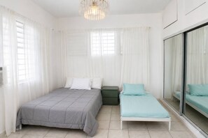 Ein Schlafzimmer mit einem großen Bett, einem kleineren Bett, einem grünen Nachttisch und einem großen Fenster mit Vorhängen.