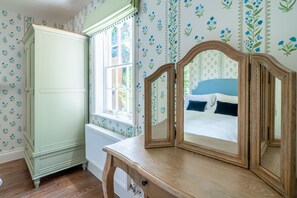 Ein Zimmer mit einem Holzkoffer, einem Spiegel, einem Fenster mit Blick auf ein Bett und blumengemustertem Wandpapier.